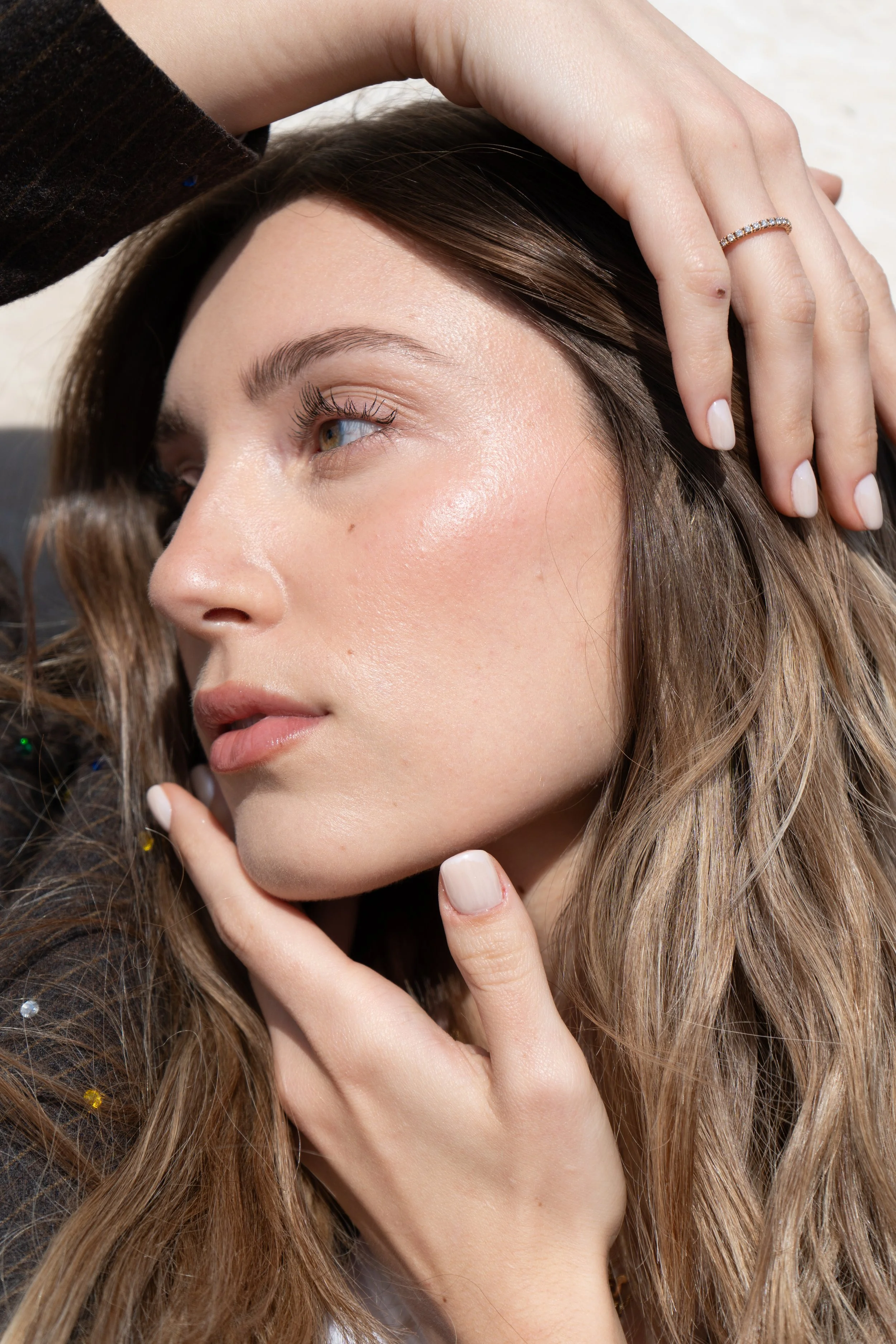 Primo piano di una donna con capelli castani ondulati, che tocca i capelli con una mano e indossa un anello con pietre preziose. La sua pelle è luminosa, con trucco leggero e sopracciglia ben definite.