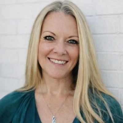 A smiling woman with long blonde hair wearing a teal top standing in front of a white brick wall.