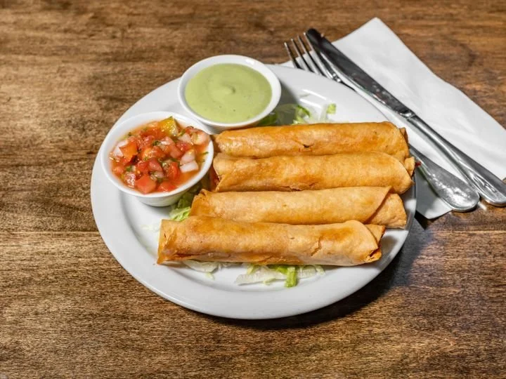 Plate of four chimi rolls with side of pico de gallo and green dipping sauce on a white plate, set on a wooden table with silverware and napkin.
