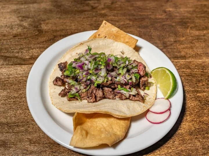 Tacos de carne asada with chopped onions and cilantro, lime wedge, radish slices, and a side of fried plantain on a white plate.