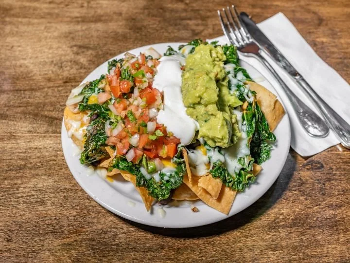 Plate of nachos topped with salsa, sour cream, guacamole, melted cheese, and cooked greens, served with a fork and knife on a napkin.