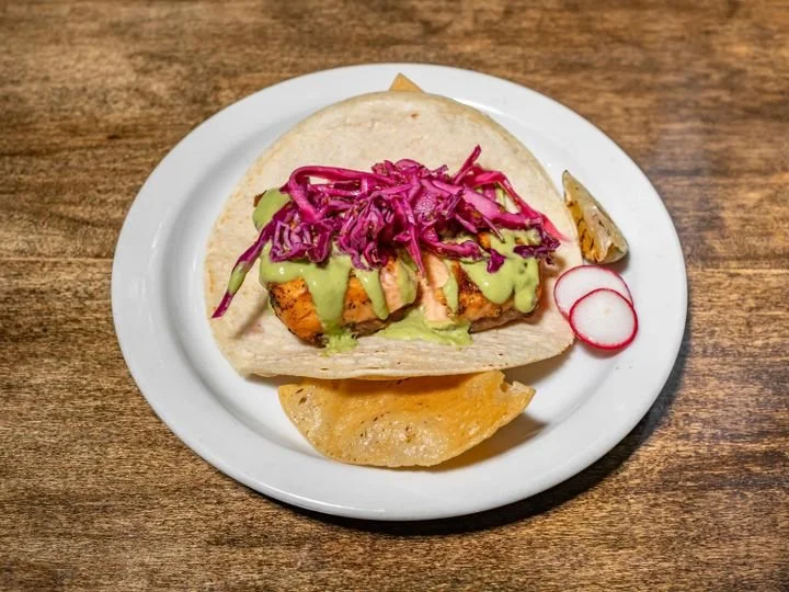 A taco with grilled chicken, shredded purple cabbage, and green sauce in a soft corn tortilla, served on a white plate with radish slices and a lemon wedge on the side.