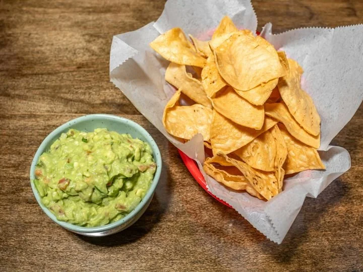 Bowl of guacamole and a basket of tortilla chips on a wooden table.