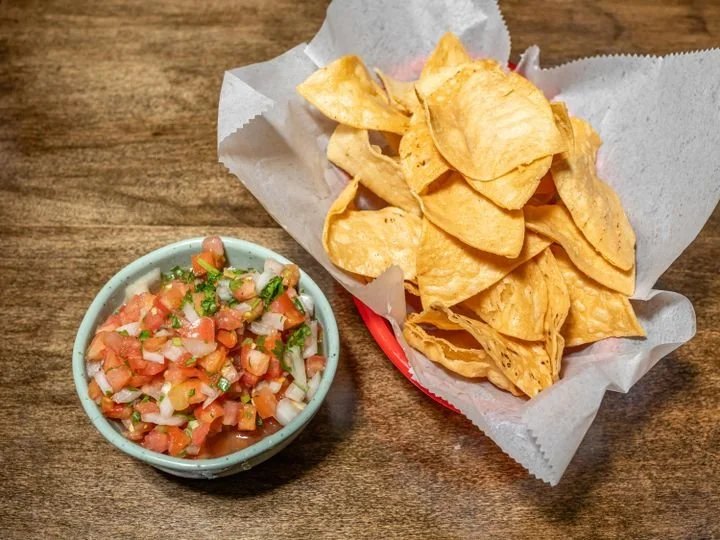 A bowl of pico de gallo and a basket of potato chips on a wooden table.
