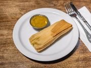 Dry tamales on a white plate with a small dish of yellow sauce, placed on a wooden table with a fork and knife.