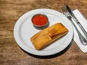 A white plate with a small bowl of ketchup and a folded egg roll on a wooden table with a fork and knife beside it.
