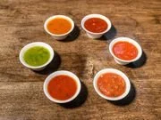 Six small bowls of various colored spices or powders on a wooden surface.