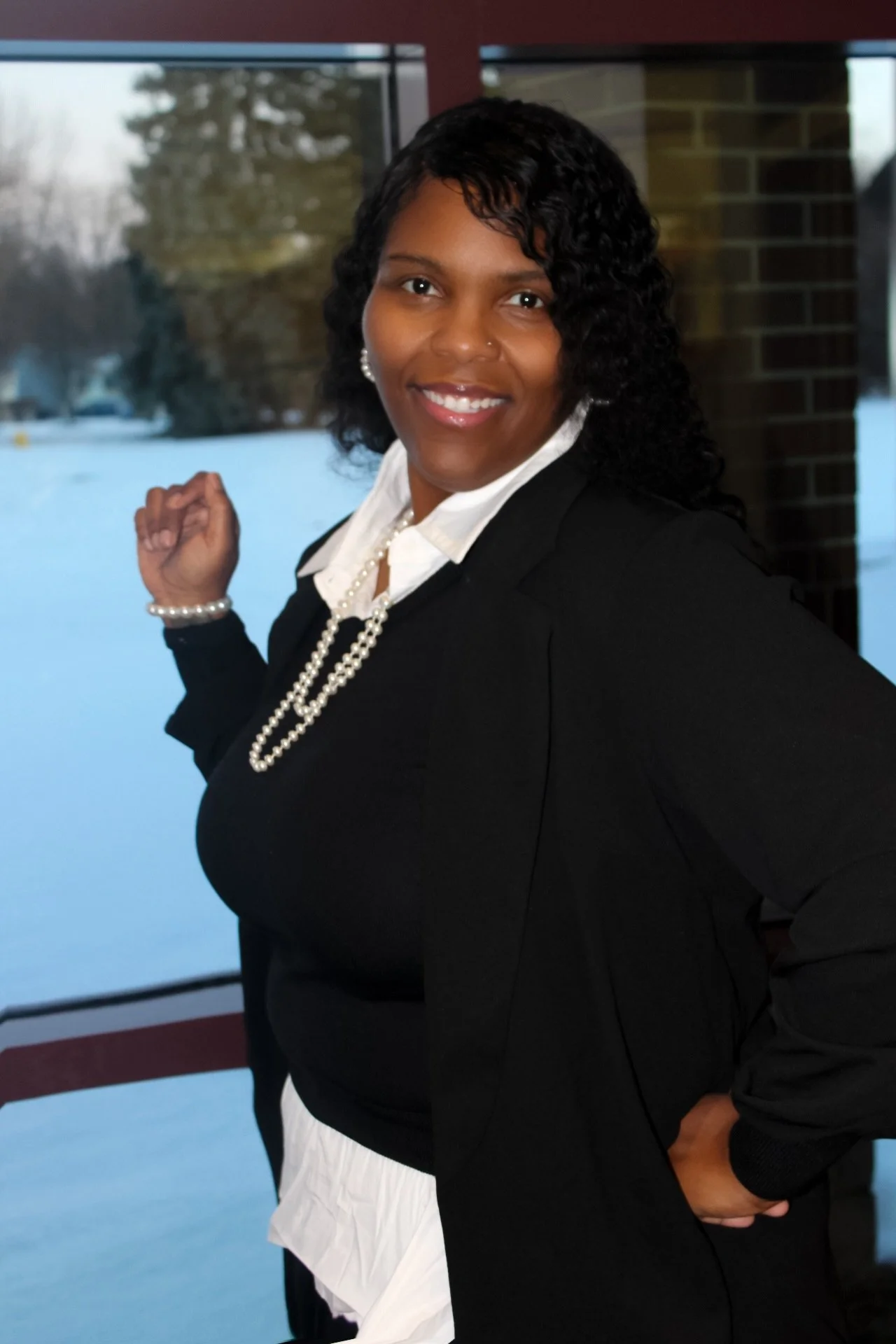 A woman with curly black hair, wearing a black blazer, white shirt, pearl necklace, and pearl bracelet, standing indoors with a snowy landscape outside the window behind her, smiling at the camera.