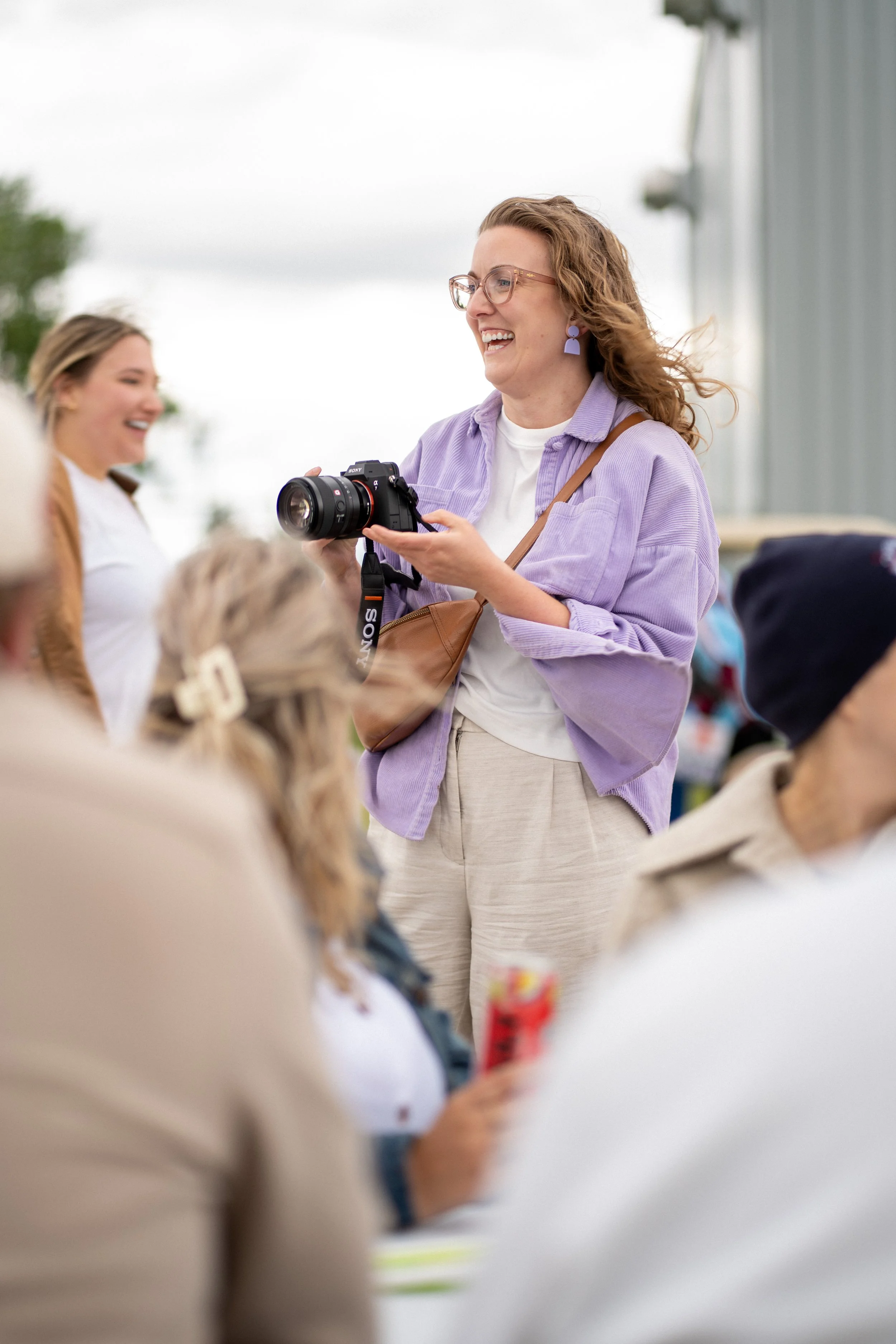 A woman with curly hair, glasses, and purple earrings smiling while holding a camera, surrounded by children, outdoors under cloudy sky.