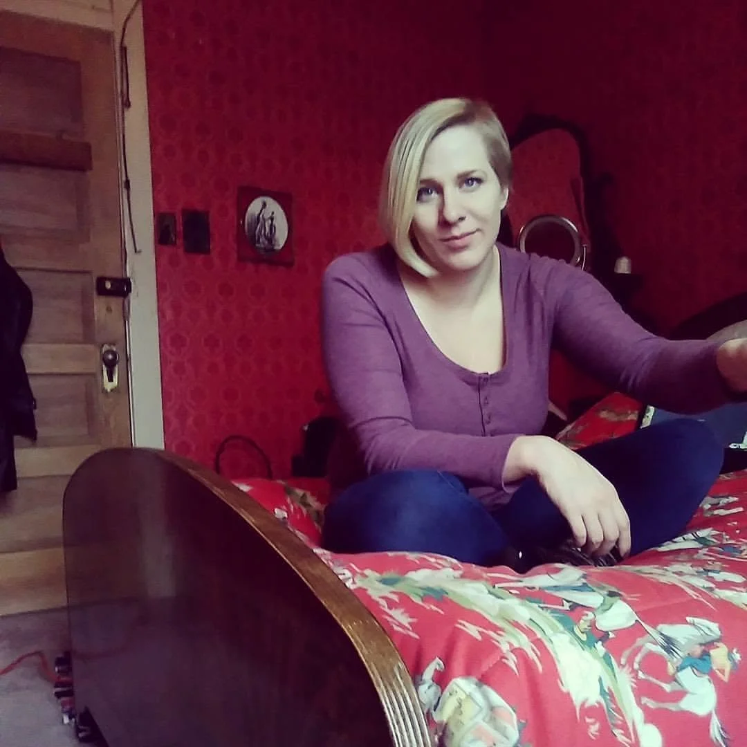 A woman with blonde hair wearing a purple long sleeve shirt, sitting cross-legged on a bed with a red holiday-themed coverlet. She is looking at the camera in a room with red patterned wallpaper and a mirror in the background.