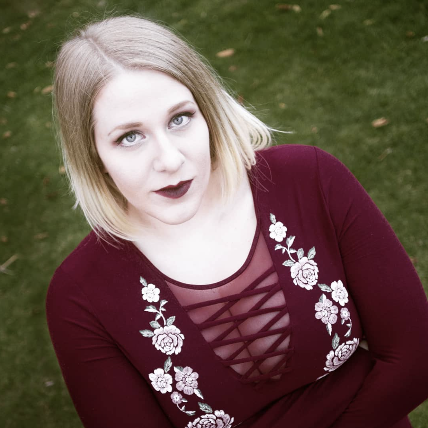 A woman with blonde hair and blue eyes wearing a burgundy dress with floral embroidery and crisscross lace-up design on the chest, standing outdoors on grass.