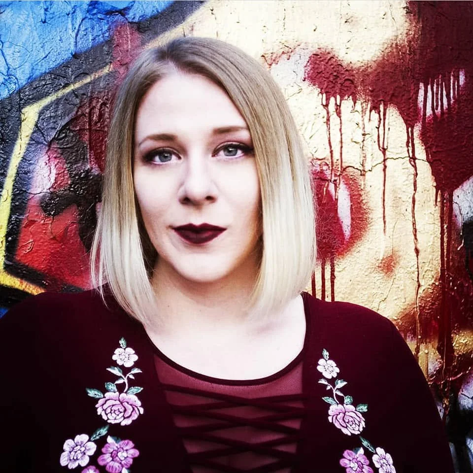 A woman with blonde hair and dark lipstick standing in front of a colorful graffiti wall.