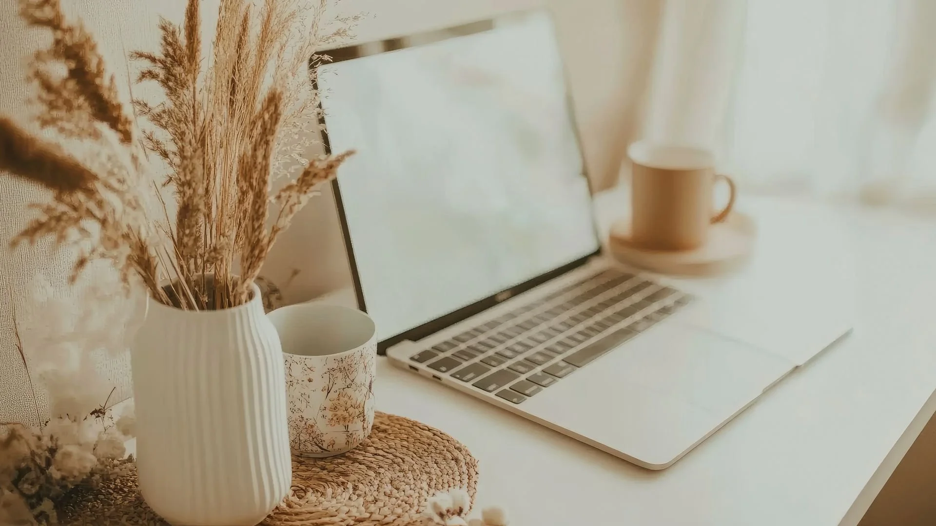 A desk with a white laptop, a mug, and a vase of dried flowers, with soft lighting and neutral tones.