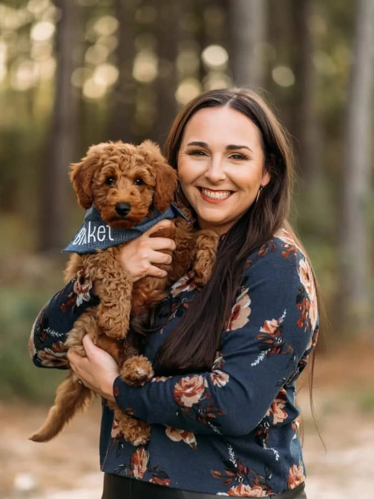 A woman with long dark hair wearing a floral jacket, smiling, holding a small brown poodle puppy with curly fur, outdoors in a wooded area.