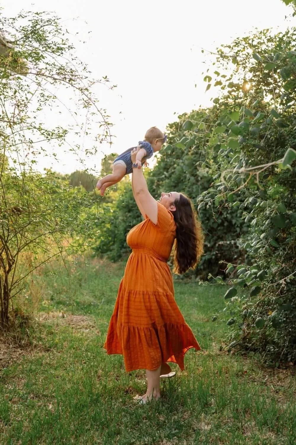 A woman wearing an orange dress lifts a baby into the air on a grassy path lined with green bushes and trees, with sunlight peeking through the leaves.