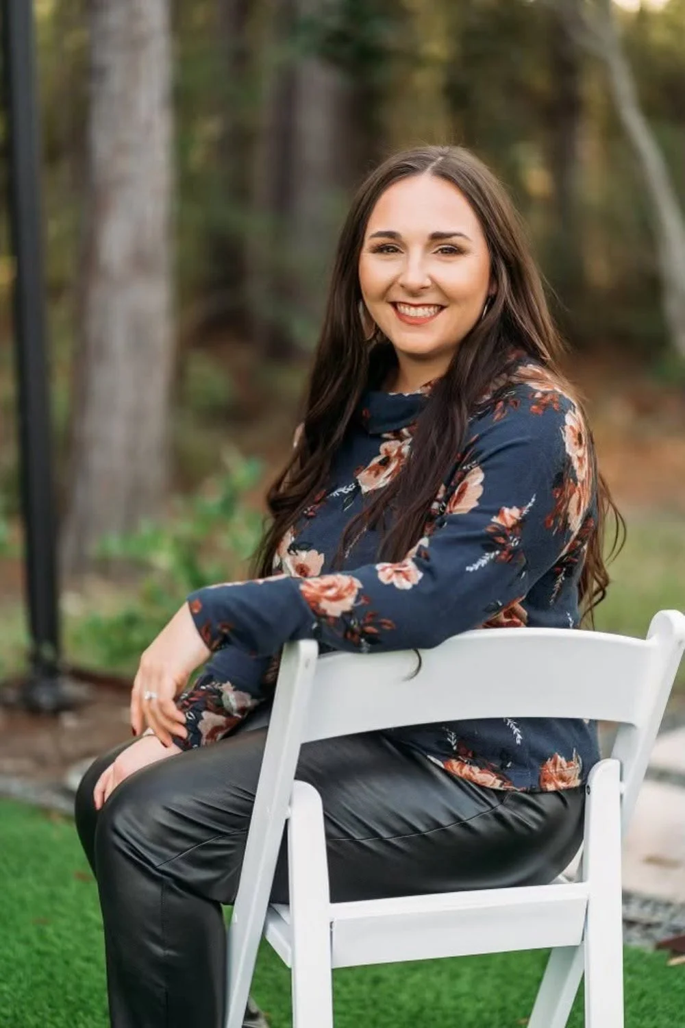 A woman with long brown hair, smiling, sitting on a white chair outdoors in a wooded area with trees in the background, wearing a floral dark blue blouse and black pants.