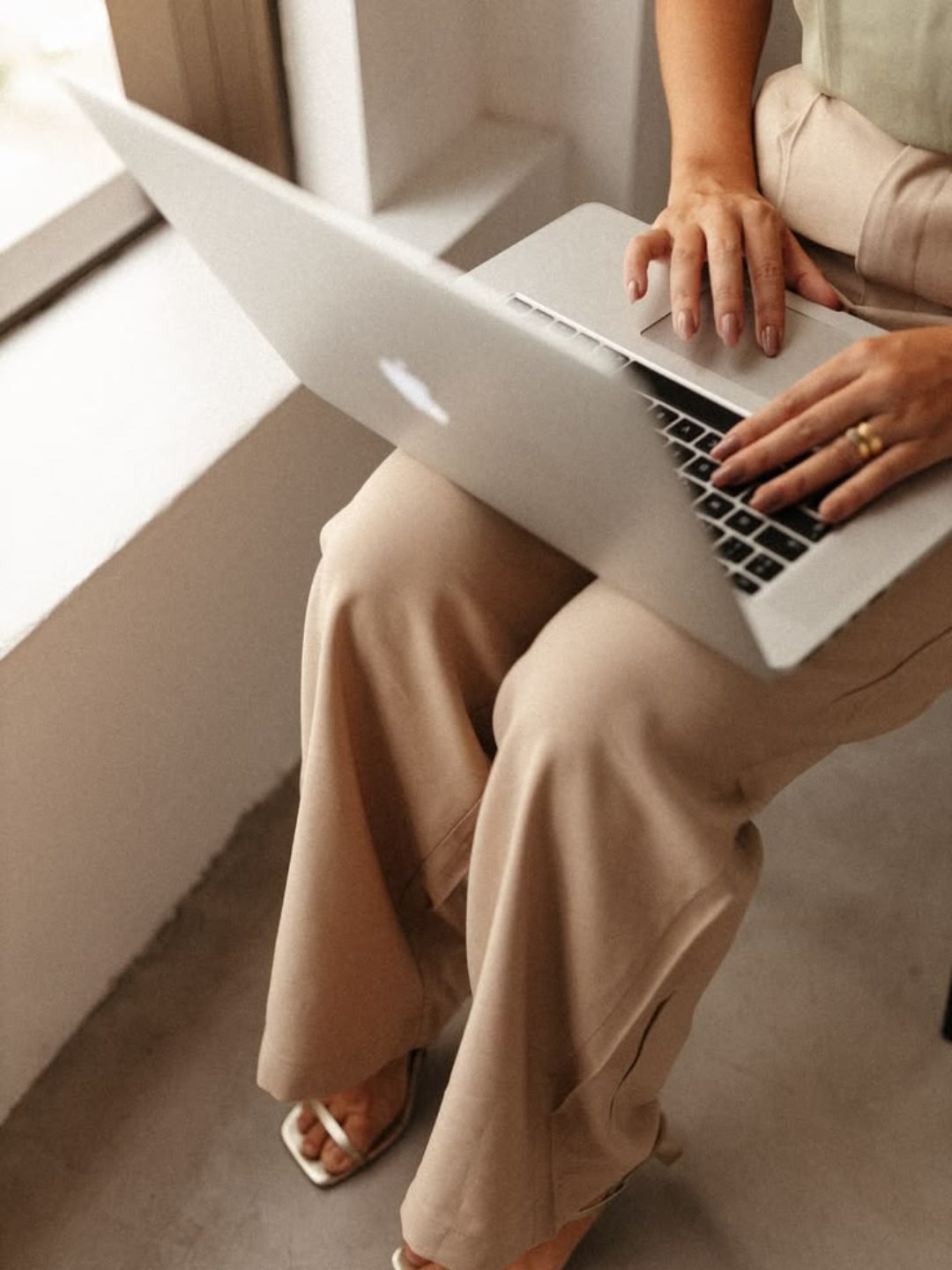 Person sitting on a ledge working on a silver MacBook laptop, wearing beige pants and sandals.
