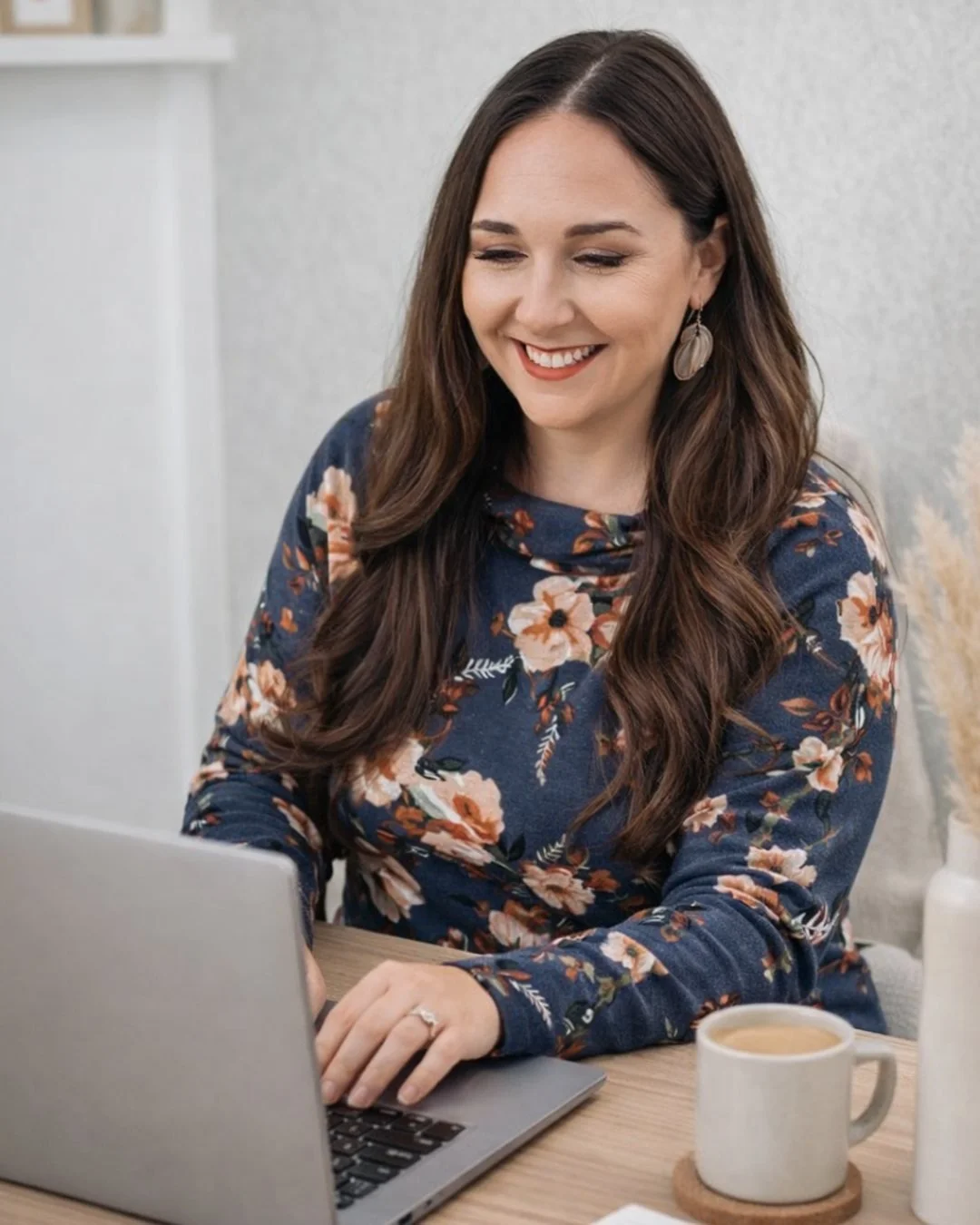 A woman with long wavy brown hair smiling while typing on a laptop at a wooden table. She is wearing a dark floral long-sleeve top, earrings, and a wedding ring. There is a mug and a white bottle on the table, and a white textured wall in the background.