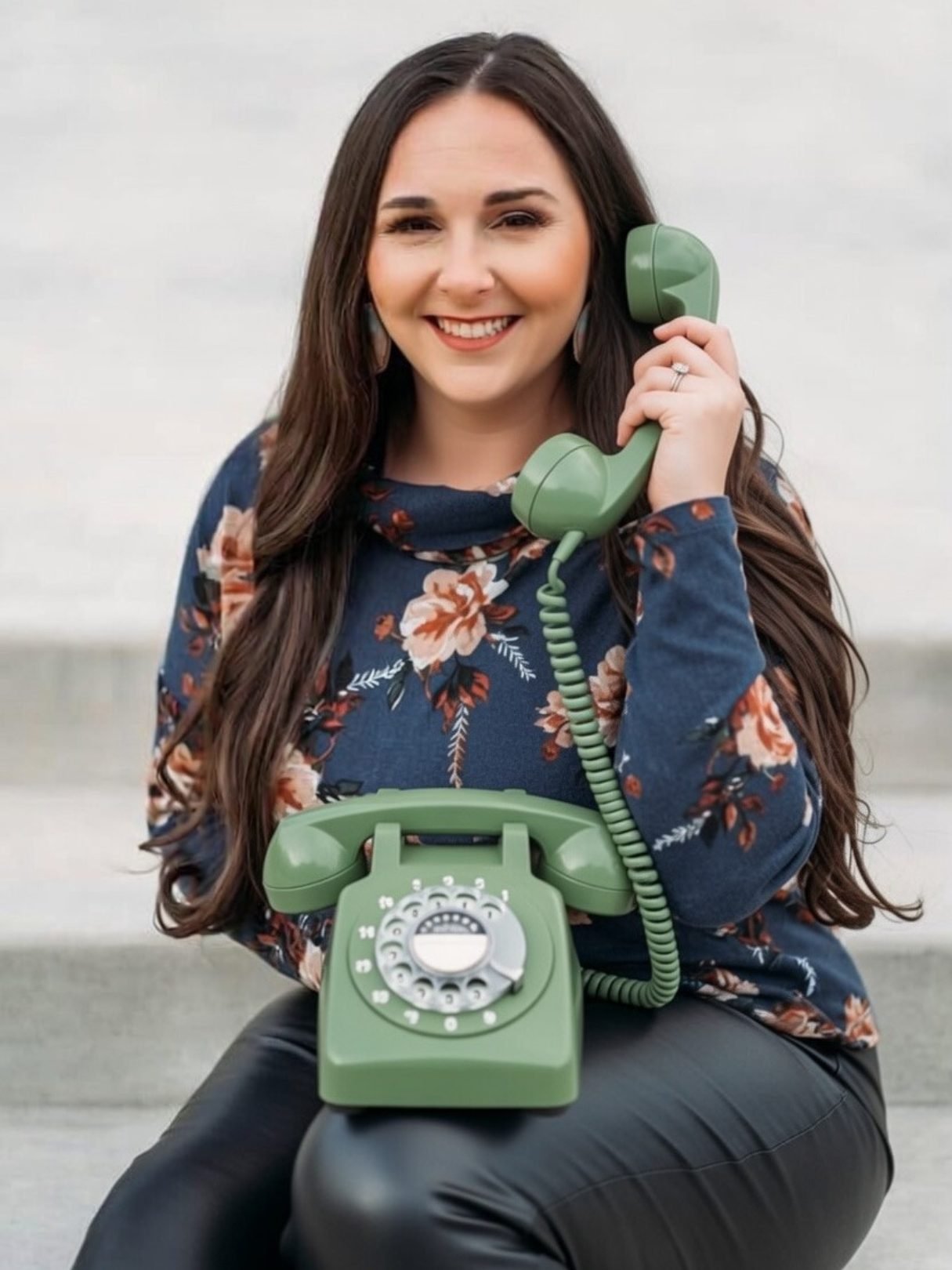 A woman with long dark hair smiling while holding a green vintage rotary phone to her ear, sitting outdoors.