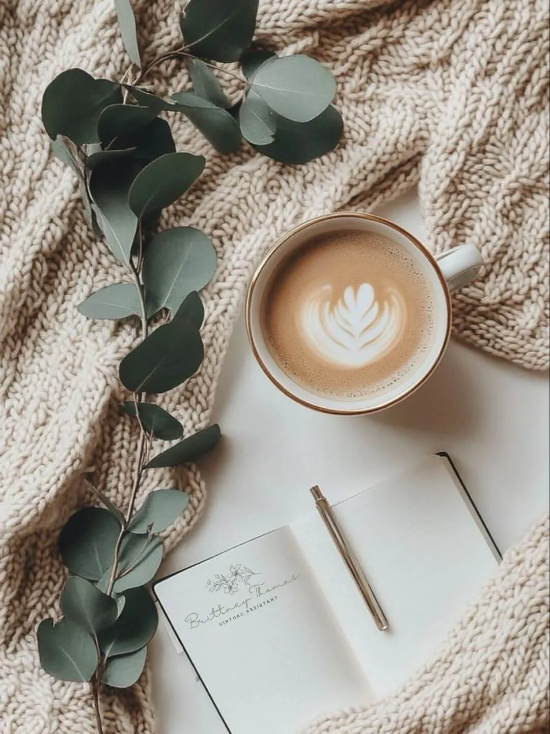 A cozy scene with a cup of coffee featuring latte art, a sprig of eucalyptus leaves, a beige chunky knit blanket, a blank notebook with a gold pen, and the name 'Buttner Thomas Virtual Assistant' on the page.