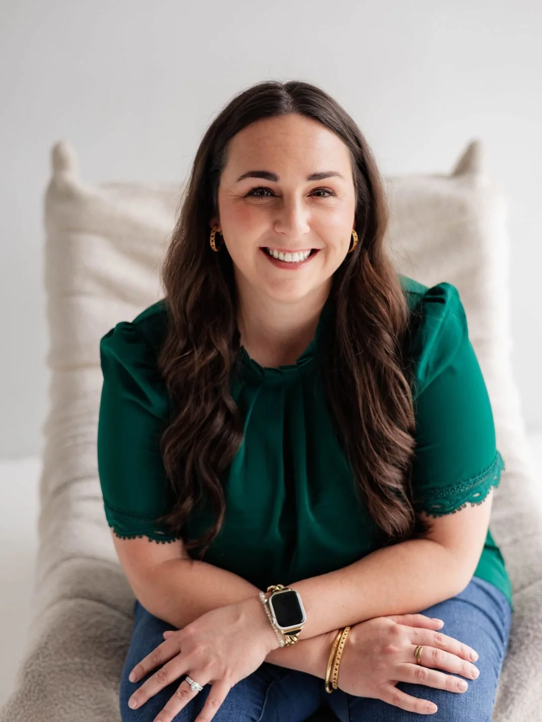 A woman with long brown hair, wearing a green top and jeans, sitting on a beige couch, smiling at the camera.