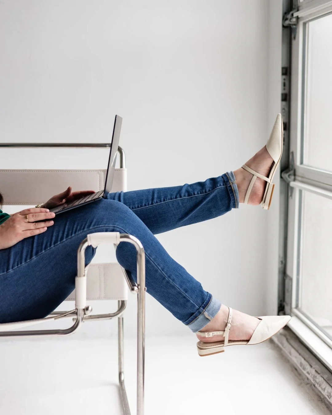 Person wearing beige pointed toe heels and blue jeans with legs extended on a chair, working on a laptop, by a large window.