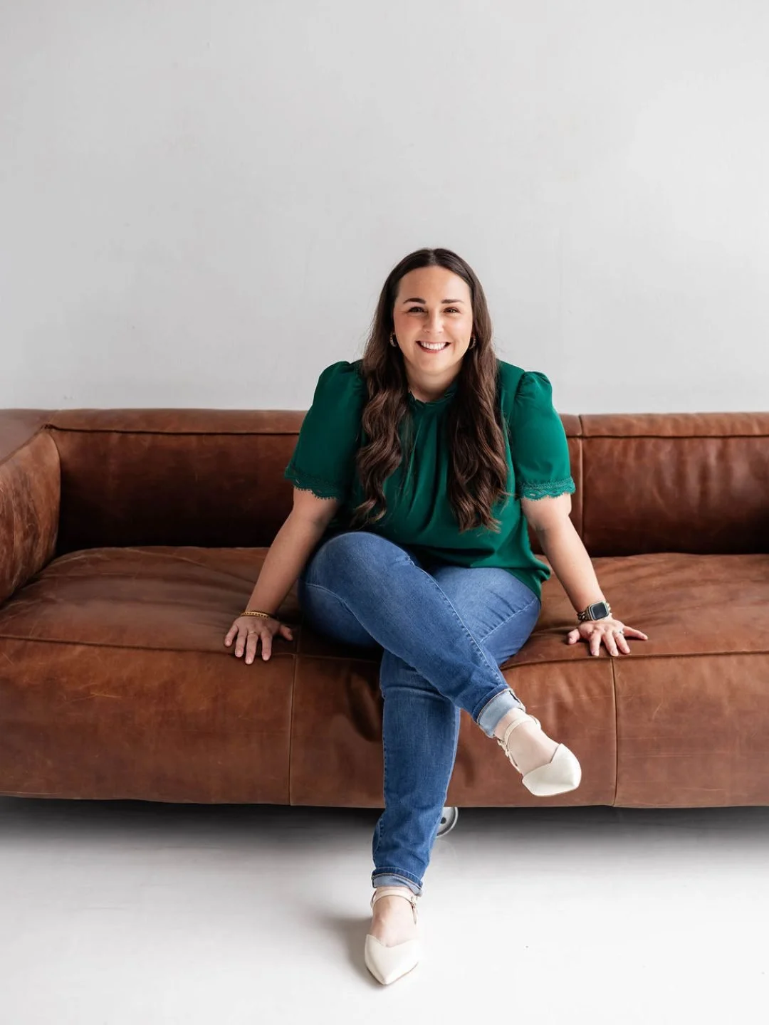 Woman sitting on a brown leather sofa, smiling, wearing a green blouse, blue jeans, and white shoes, with a white wall background.