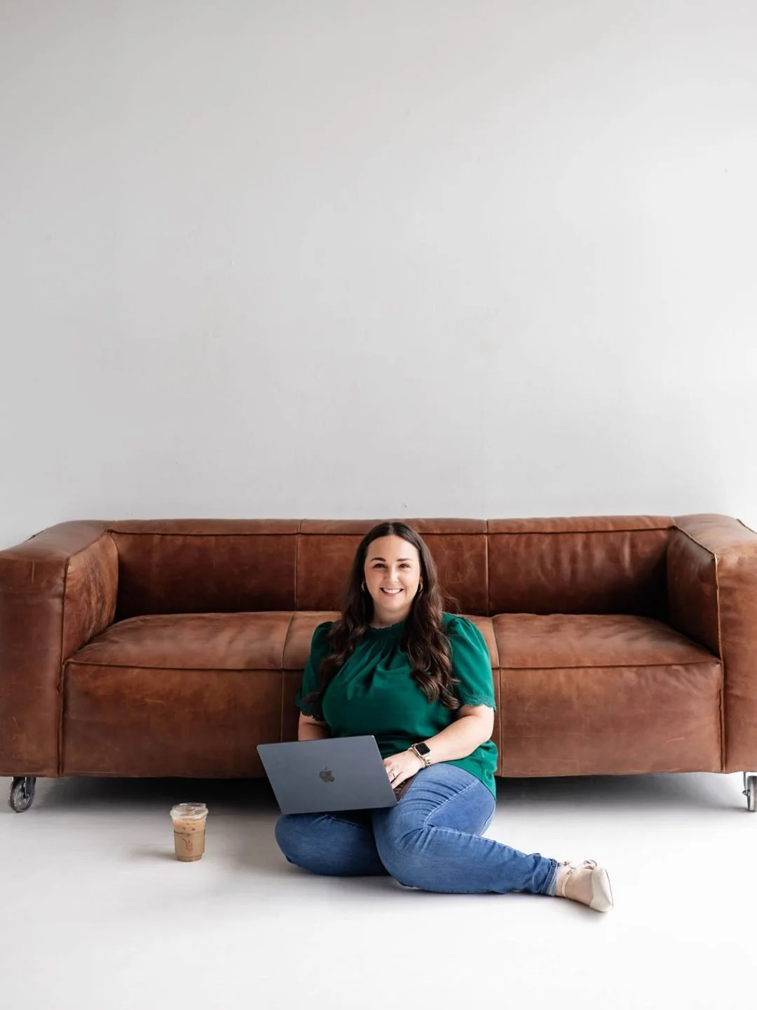 A woman with long dark hair, wearing a green blouse, blue jeans, and white shoes, sitting on the floor with a laptop on her lap, smiling at the camera, next to a brown leather sofa and a glass of iced coffee.