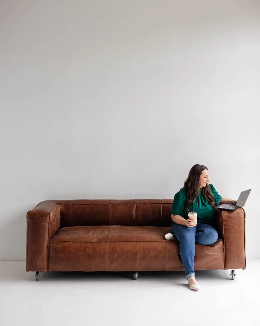 Woman sitting on a brown leather sofa, holding a coffee and using a laptop, against a plain white wall.