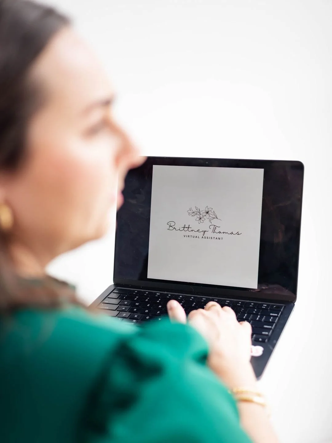 Woman viewing a laptop screen displaying a logo with floral design and the text 'Brittney Thomas Virtual Assistant'