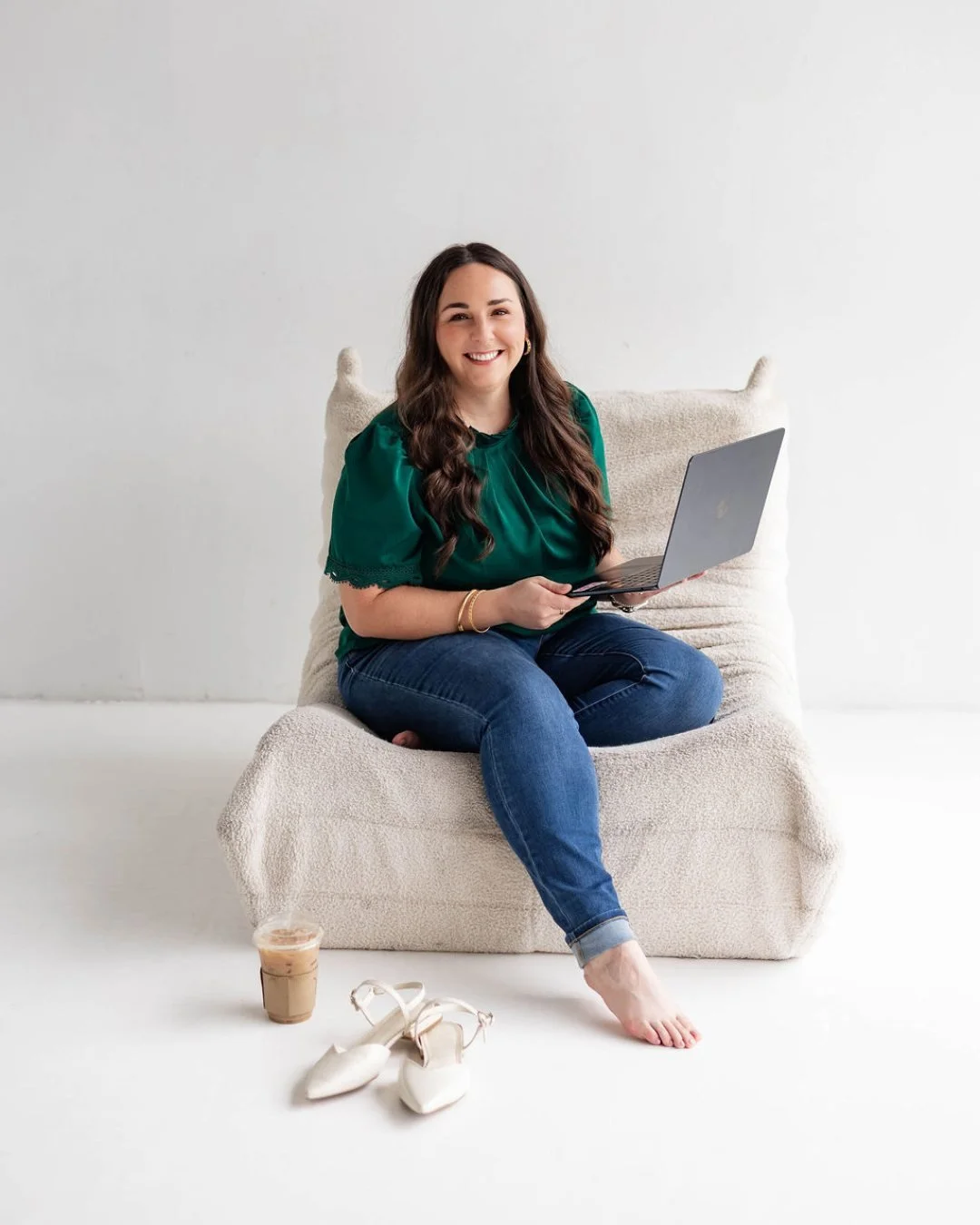 A woman with long dark hair sitting on a beige couch, holding a laptop, in a white room with a drink and white shoes on the floor.
