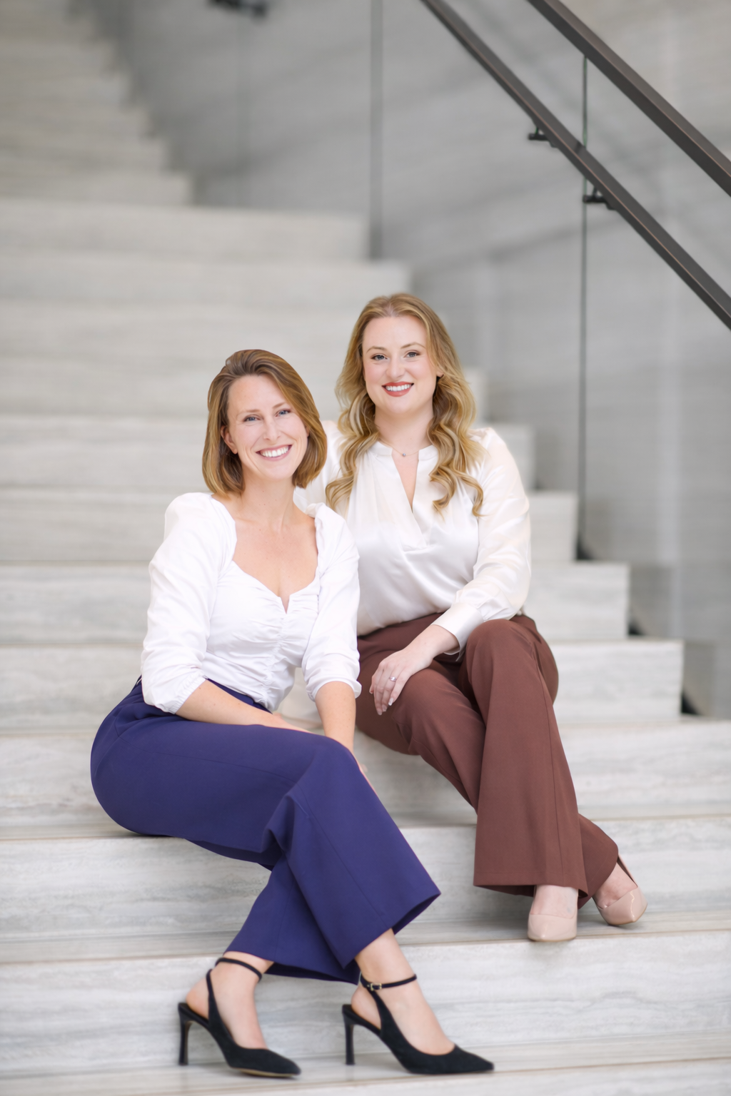 Two women sitting on concrete stairs, smiling at the camera. One with shoulder-length brown hair, wearing a white blouse and blue pants, and black heels. The other with long blonde hair, wearing a white blouse and brown pants, and beige heels.