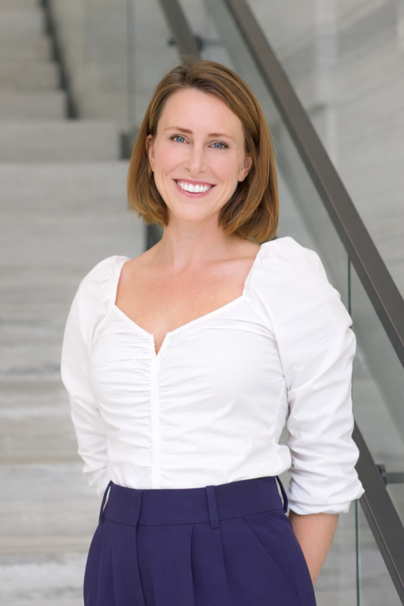 A woman with shoulder-length auburn hair, smiling, wearing a white blouse with ruched front and navy blue pants, standing indoors near a staircase.