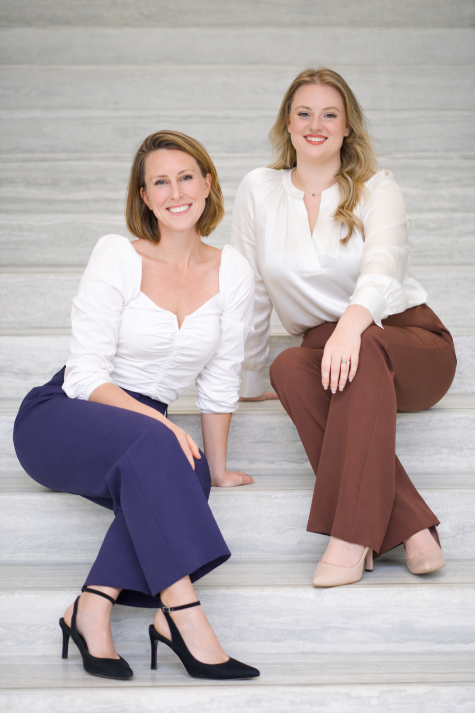 Two women sitting on marble stairs, smiling at the camera, one with short brown hair wearing a white blouse and navy pants, and the other with long blonde hair wearing a white blouse and brown pants.