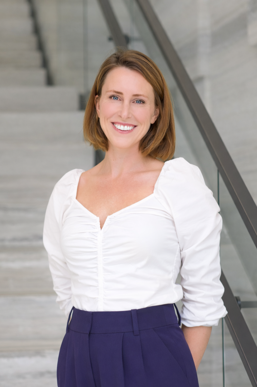 A smiling woman with shoulder-length brown hair, wearing a white blouse and dark high-waisted pants, standing indoors in front of a staircase.