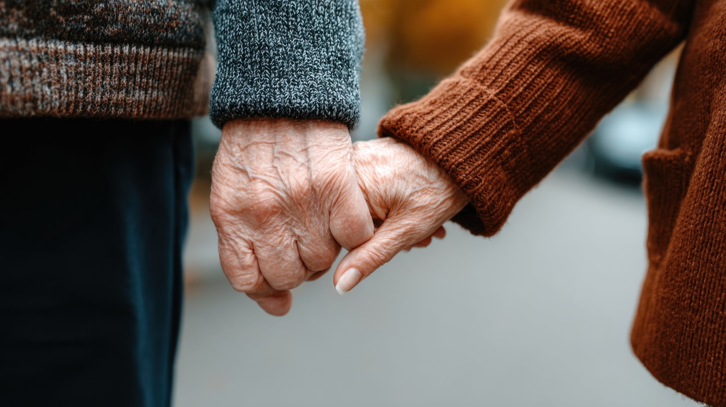 Close-up of an elderly couple holding hands, showing wrinkles and veins, with the woman finger resting on the man's finger, both wearing sweaters.