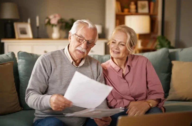 An elderly man and woman sitting on a couch looking at papers together, in a cozy living room.