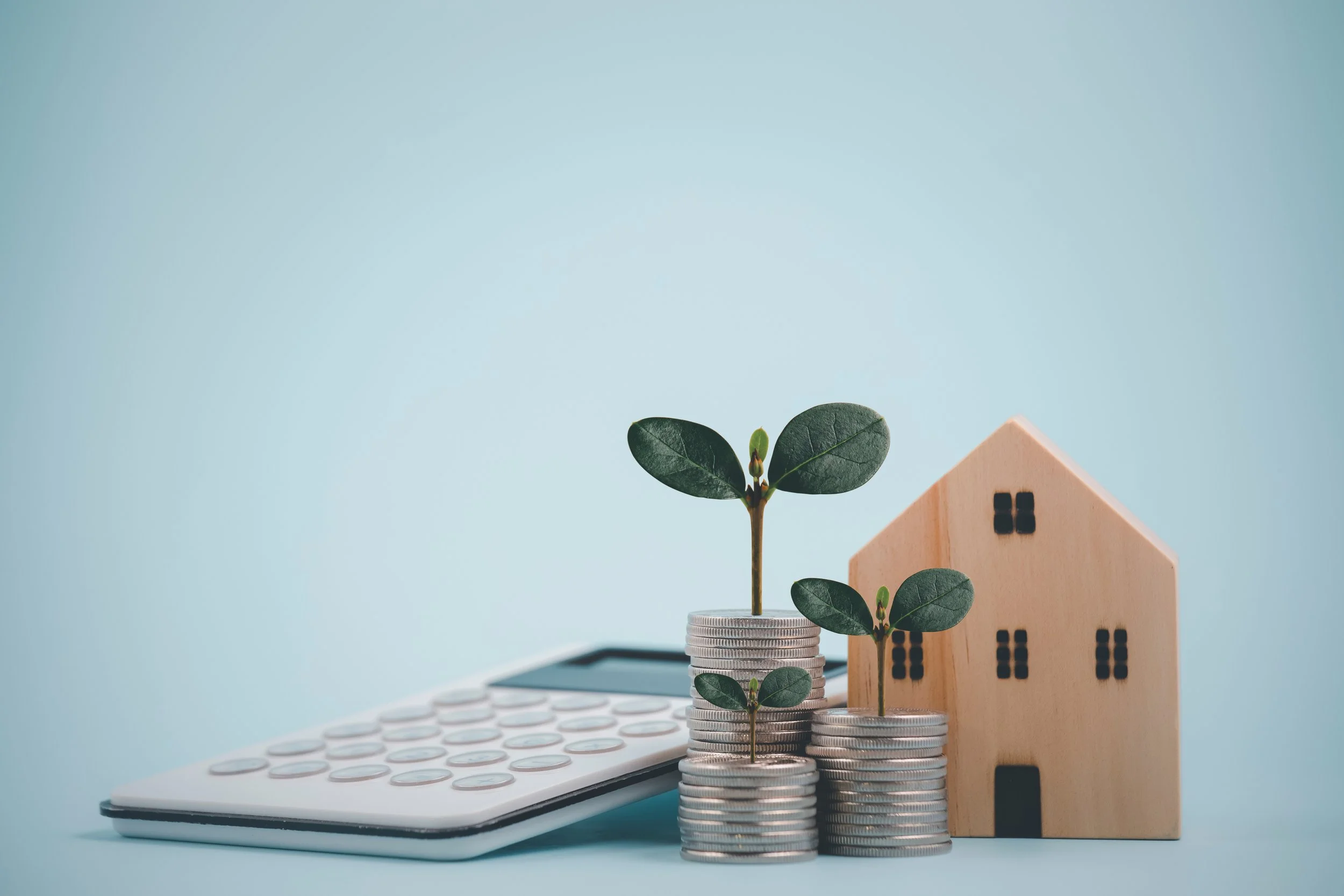 Coins stacked with small plants growing on top, a wooden house model, and a calculator placed on a light blue surface.