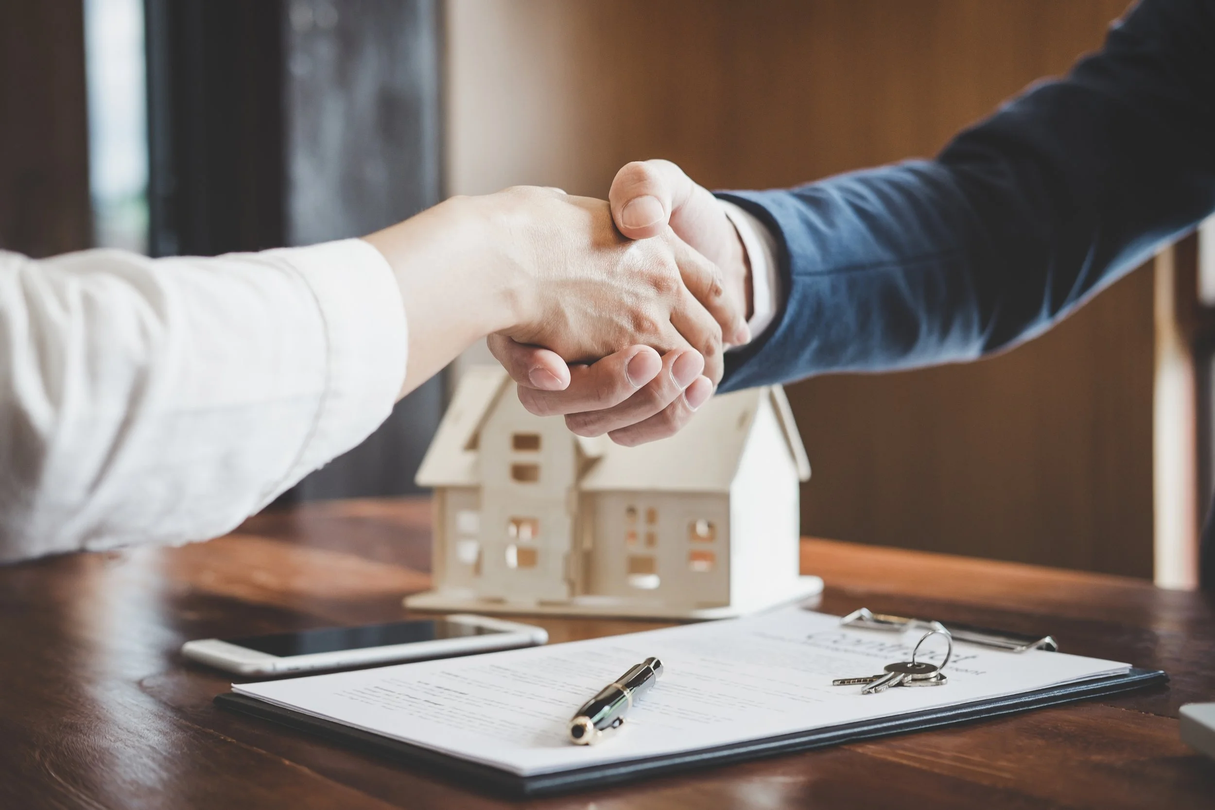 Close-up of two people shaking hands over a table with a toy house, a clipboard with documents, a pen, keys, and a phone, suggesting a real estate agreement or contract signing.