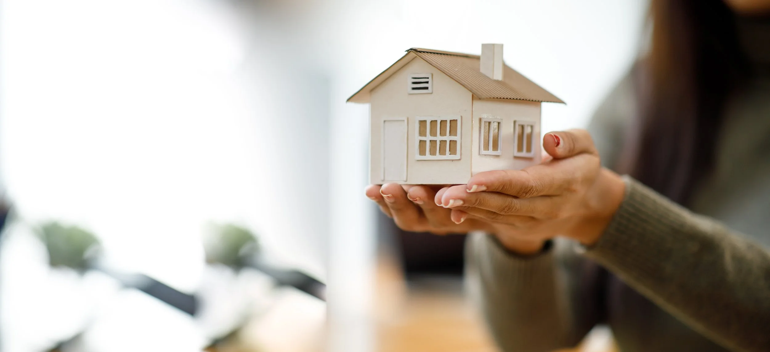 Person holding a small model house with both hands in a bright indoor setting.