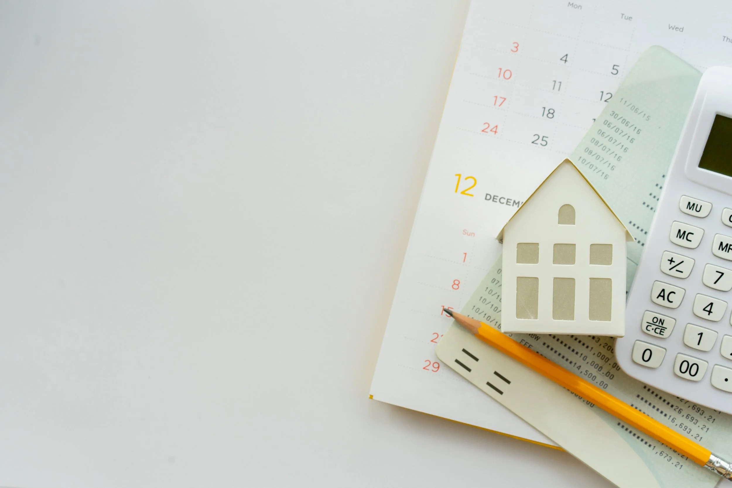 A flat lay of a calculator, a small house-shaped model, a pencil, a bank statement, and a calendar open to December, all on a white surface.