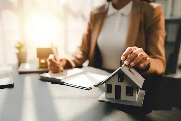 Person in business attire holding a small model house on a desk in an office setting.