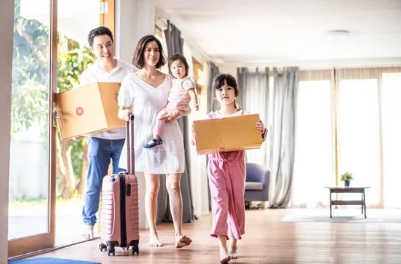 Family of four walking into a home with boxes, a woman holding a toddler, a young girl carrying a box, and a man with a suitcase, indicating moving in.