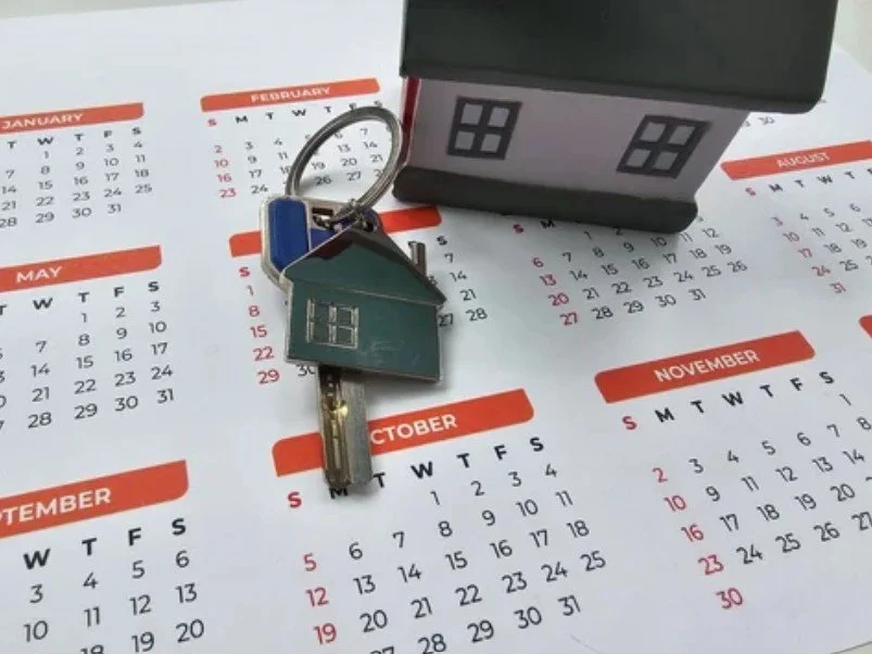 Small house-shaped keychain with house and window design placed on an open calendar, with a house-shaped object behind on the calendar pages.