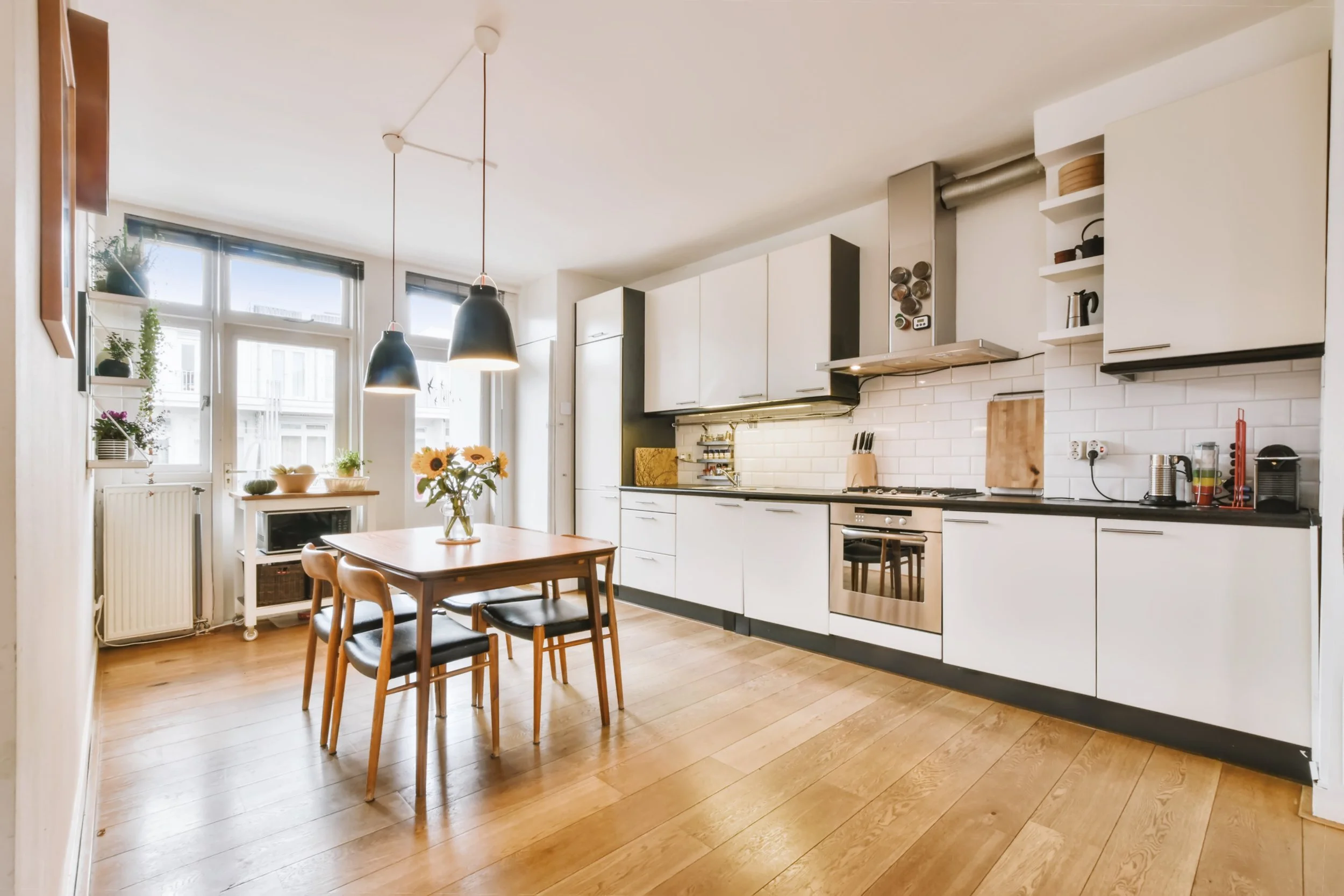 Modern kitchen with white cabinets, black countertops, and wooden flooring. There is a wooden dining table with four chairs, a vase with sunflowers, and various kitchen appliances and utensils.