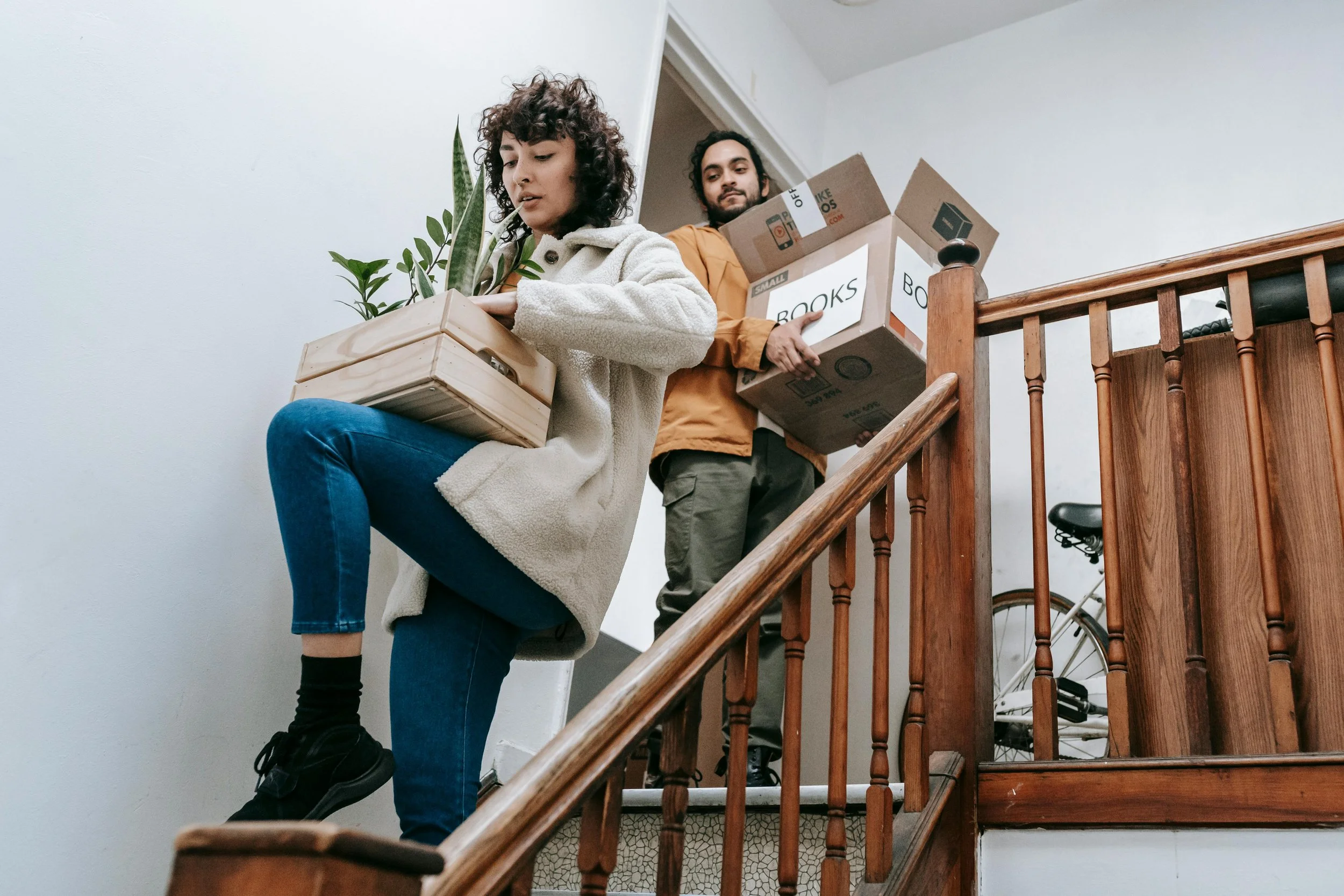 Two people carrying boxes and a plant down a staircase inside a home.