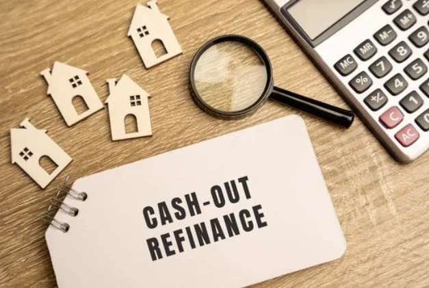 A notebook with 'Cash-Out Refinance' written on it, surrounded by small white house cutouts, a magnifying glass, and a calculator on a wooden surface.