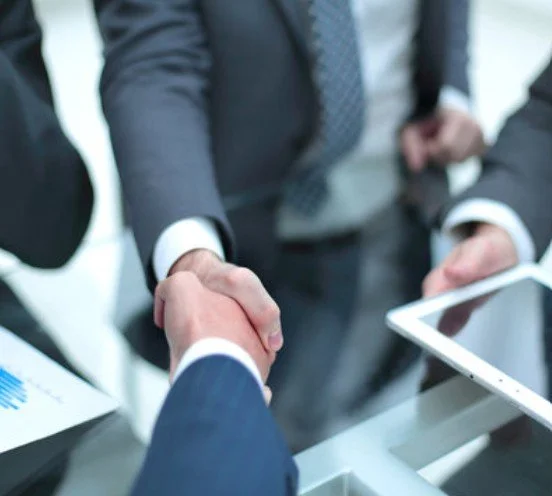 Two people in business suits shaking hands over a desk in an office setting.