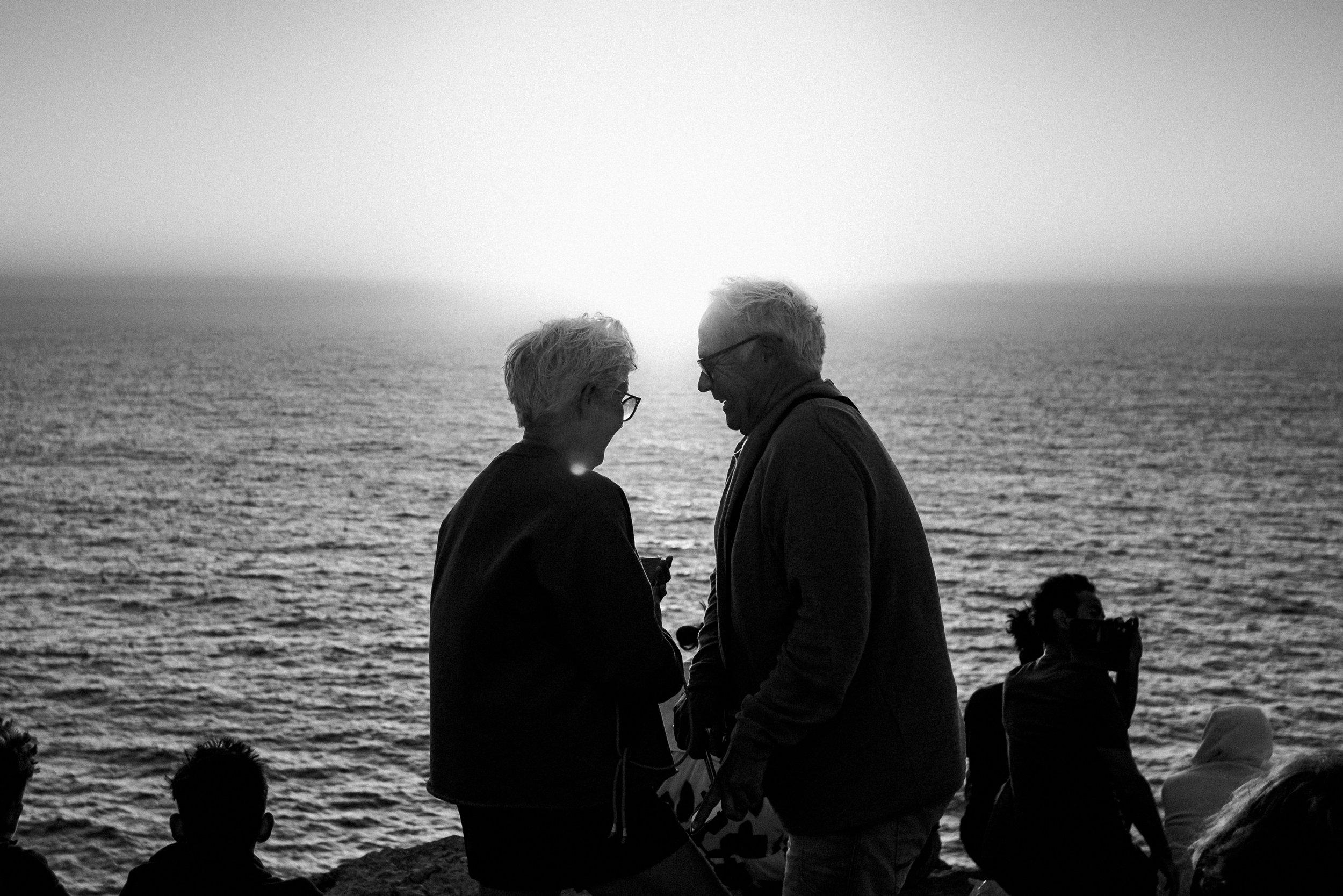 Black and white photo of an older couple smiling and looking at each other on a beach at sunset, with the ocean in the background, and a few other people sitting or standing nearby.