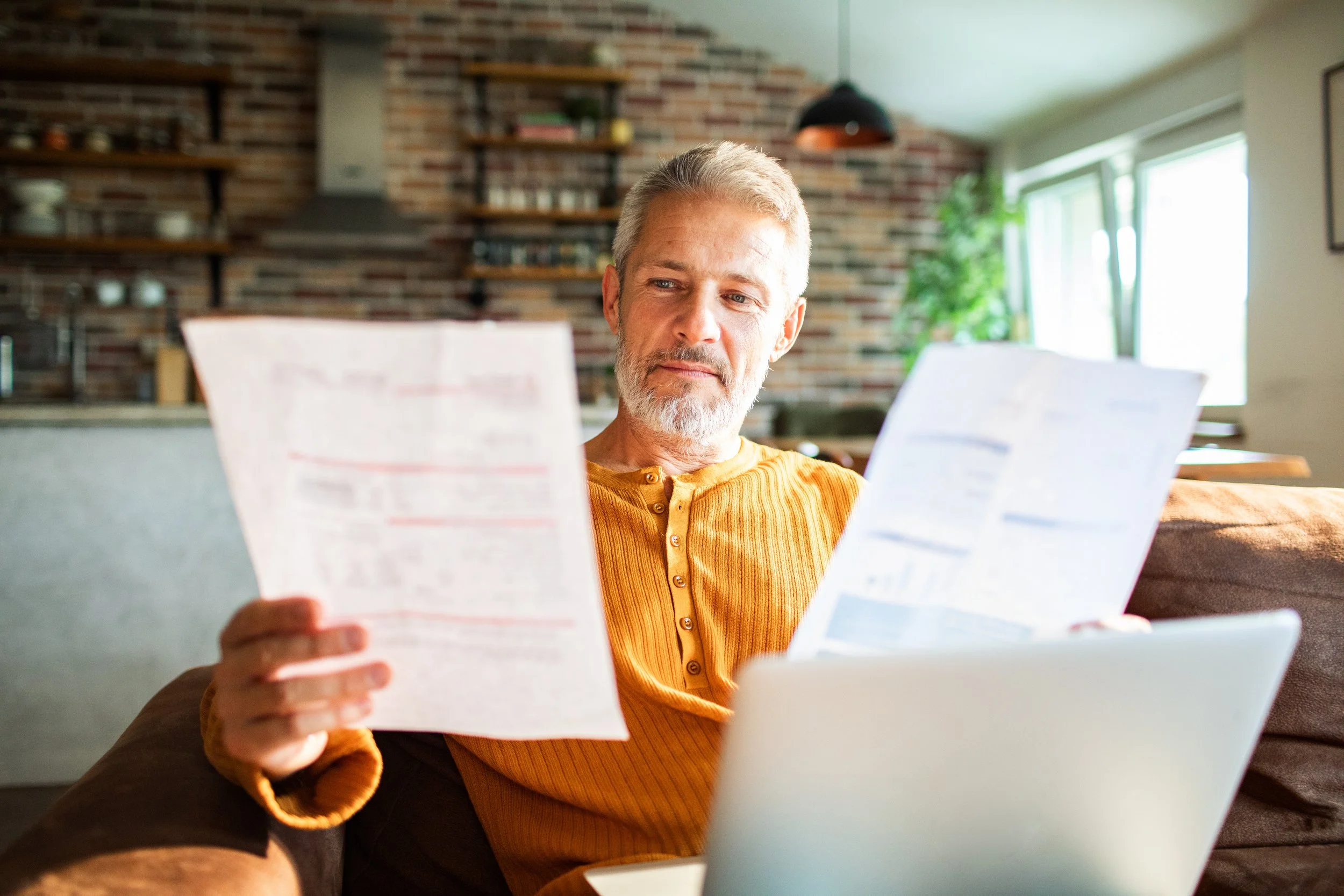An older man with gray hair and beard sitting on a couch, reading papers while using a laptop in a bright, modern kitchen.